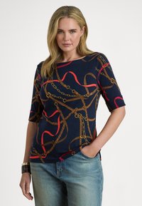 Žena s blond vlasy, oblečená v tmavě modrém topu s červeným a hnědým vzorem řetězů a pásků, modré džíny a tmavý náramek, stojící s jednou rukou v kapse.