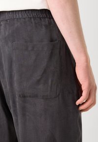 Pantalons gris foncé en tissu lisse, présentant une taille élastique et une poche arrière unique avec des détails de couture soignés.