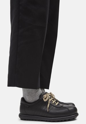 Persona con pantalones negros, calcetines grises y zapatos de cuero negro con cordones beige sobre un fondo blanco.