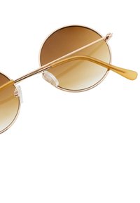 Pyöreät aurinkolasit, joissa on sävyttyneet ruskeat linssit, kultasävyinen metallikehys ja läpinäkyvät silikoniset nenätuet. Kestäviä kaarevia templeitä, joissa on yksityiskohtia.