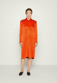 Vestido de manga longa e com botões em cetim laranja vibrante, com colarinho e um hem ligeiramente curvado. Combinado com sapatos pretos baixos.