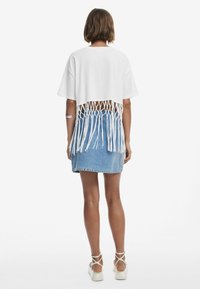 Biely skrátený top s dizajnom strapcov na chrbte, v kombinácii s modrou džínsovou sukňou. Top má voľný strih a štruktúrovaný zadný detail.