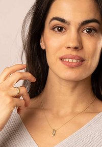 Goldene Halskette mit einem rechteckigen Anhänger, der drei kleine Edelsteine zeigt. Die Kette wird von einer Person mit schulterlangem dunklem Haar und einem hellgrauen Oberteil getragen.