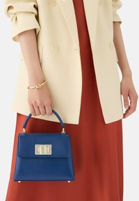 Blauwe gestructureerde handtas van textuurleer, met een bovenhandvat en een metalen sluiting. Draagt samen met een crèmekleurige blazer en een rode jurk.