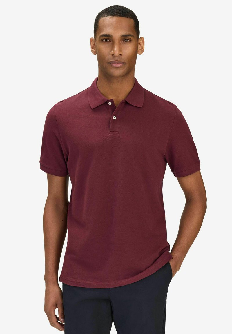 Homme portant un polo manches courtes couleur bordeaux et un pantalon sombre, debout avec une main dans la poche devant un fond blanc.