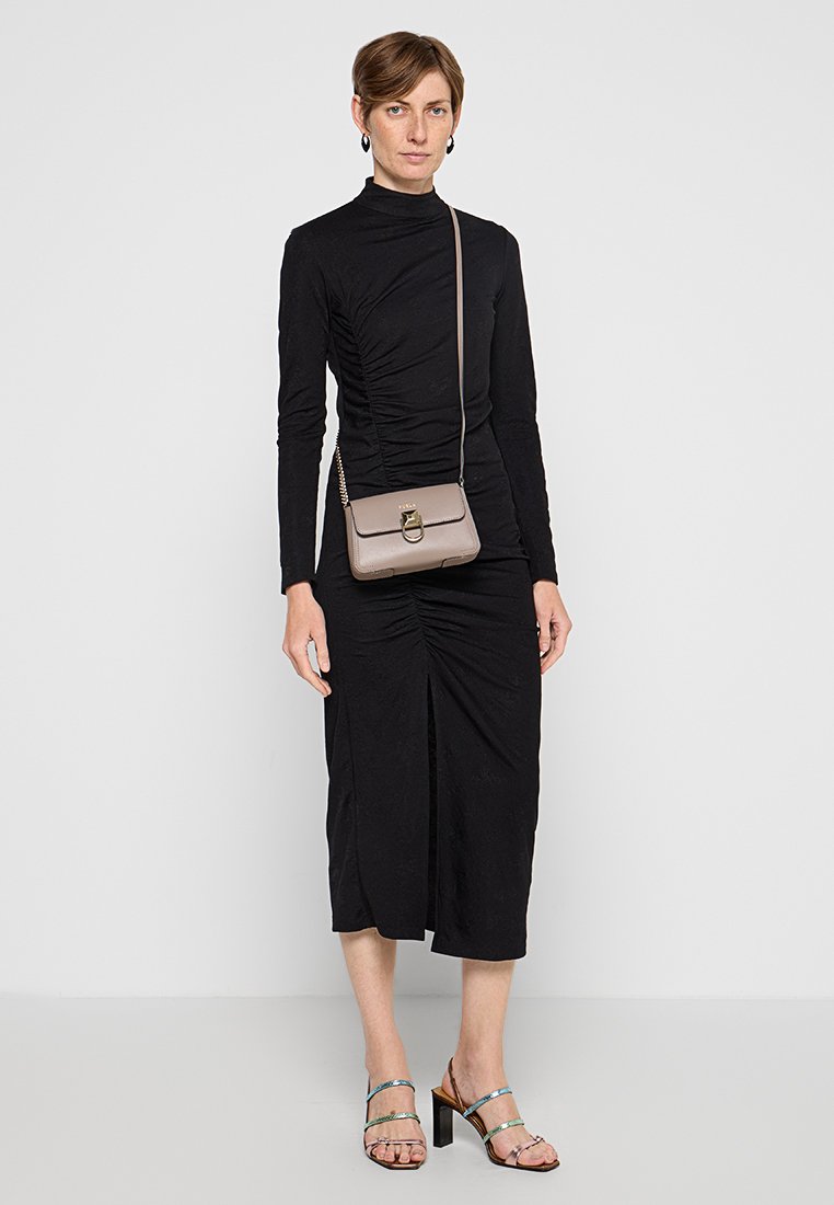 Schwarzes Kleid mit langem Ärmel und geraffter Textur, in Knielänge, kombiniert mit einer hellbraunen Umhängetasche und metallischen Sandalen mit Absatz.