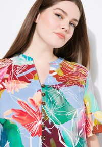 Blusa floral en azul, con diseños multicolores en acuarela. Tiene un cuello sin solapas, mangas cortas y cinco botones marrones en la parte delantera.