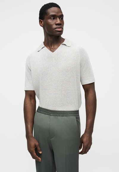 Ανοιχτό γκρι πλεκτό πόλο μπλουζάκι με κοντά μανίκια, V-λαιμόκοψη και ελαφρώς διπλωμένο γιακά, συνδυασμένο με πράσινο παντελόνι με ελαστική μέση.
