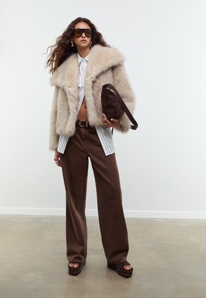 Beige Kunstpelzjacke, weiß gestreiftes Hemd, braune weit geschnittene Hose, klobige Plattformschuhe. Accessoires sind eine große Sonnenbrille und eine kleine Tasche.