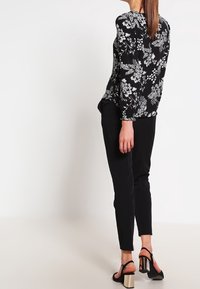 Blouse noire à fleurs avec manches longues et col rond, associée à un pantalon noir. Le modèle porte des sandales noires avec un talon métallique.