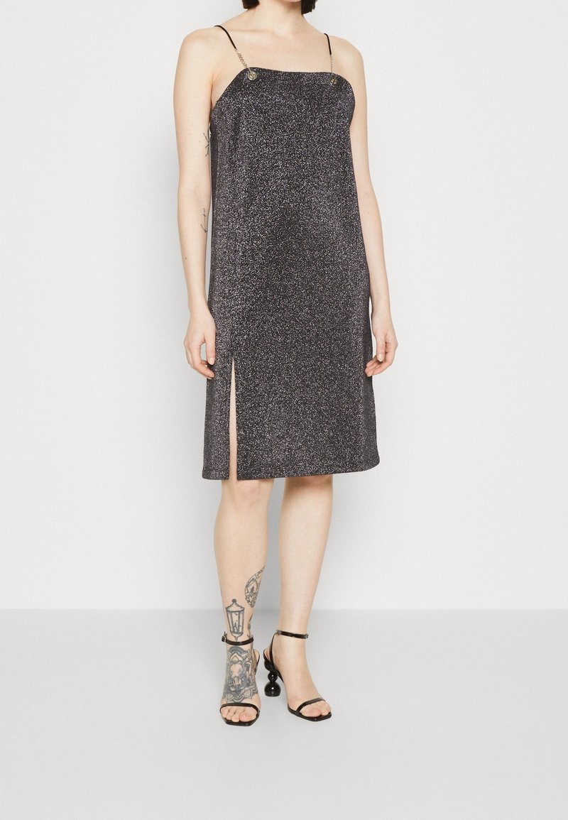 Robe sans manches longueur genou gris foncé pailleté avec fines bretelles et fente latérale, portée avec des sandales noires à talons hauts sur des jambes tatouées.