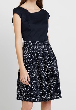 Vrouw die een marineblauwe jurk draagt tot op de knie met een effen aansluitend bovenstuk en een geplooide rok met witte stippen.