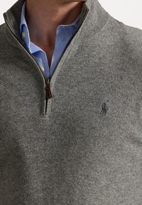 Pull gris à fermeture éclair en tissu tricoté, avec un col montant et un petit logo brodé sur la poitrine, porté par-dessus une chemise bleu clair.