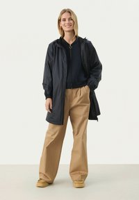 Lång svart regnkappa med huva, elastiska ärmslut och dragkedja. Matcher med lösa beige byxor och slip-on ljusbruna skor. Ljus bakgrund.