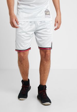 Mand iført hvide mesh basketballshorts og sorte høje sportssko står på en ensfarvet lys gulv- og vægbaggrund.