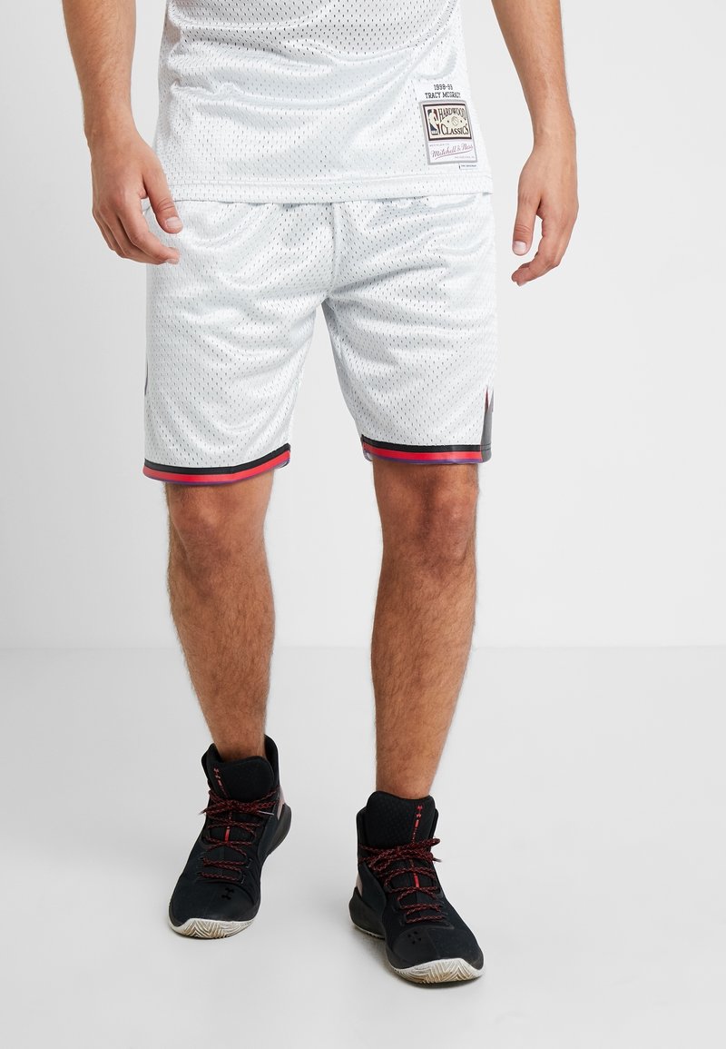 Homme portant un short de basket blanc en mesh et des chaussures de sport montantes noires, debout sur un sol et un mur clairs et sobres.