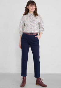 Bloes met bloemenpatroon in het wit met rode en blauwe bloemen. Marineblauwe geklede broek met zijzakken. Bruine leren enkellaarsjes.