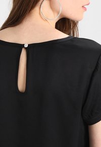 Černý krátký rukáv top s malým otvorem na zádech, který je zabezpečen knoflíkem. Tkanina se zdá být hladká a lehká.