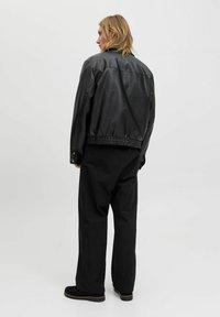 Persona rubia de pie mirando hacia la izquierda, vestida con una chaqueta de cuero negra, pantalones anchos negros y zapatos negros, contra un fondo gris liso.