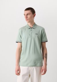 Světle zelená polo košile z bavlny, se zapínáním na knoflíky, kontrastním límečkem a černými detaily. Obsahuje logo na levé části hrudi.