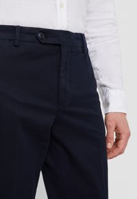 Dunkelblaue Hose mit glatter Textur, ausgestattet mit einem Knopfverschluss und Seitentaschen. Kombiniert mit einem weißen Langarmhemd.