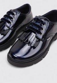 Zapatos de charol azul marino con una suela negra gruesa, que presentan un acento decorativo con flecos, seis cordones de terciopelo y ojetes plateados.