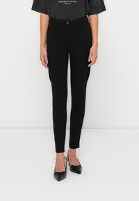 Zwarte aansluitende cargo-broek met zijzakken, gemaakt van rekbaar materiaal. Gecombineerd met zwarte puntige ballerina's. Eenvoudig en functioneel ontwerp.