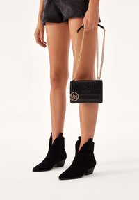 Czarna torebka typu croc z tłoczeniem, wyposażona w złotą łańcuszkową rączkę, zestawiona z czarnymi zamszowymi botkami za kostkę z ostrym noskiem i niskim słupkowym obcasem.