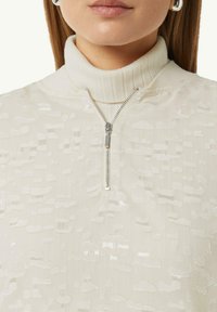 Cremfarbener Zip-Up-Pullover mit einem gerippten Kragen, der ein strukturiertes Muster aufweist und einen silbernen Reißverschlussdetail enthält, das einen subtilen Glanz verleiht.