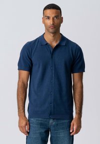 Marineblå kortærmet polo shirt lavet af strukturstof, med knaplukning foran, blød krave og ribbede manchetter, parret med denimjeans.