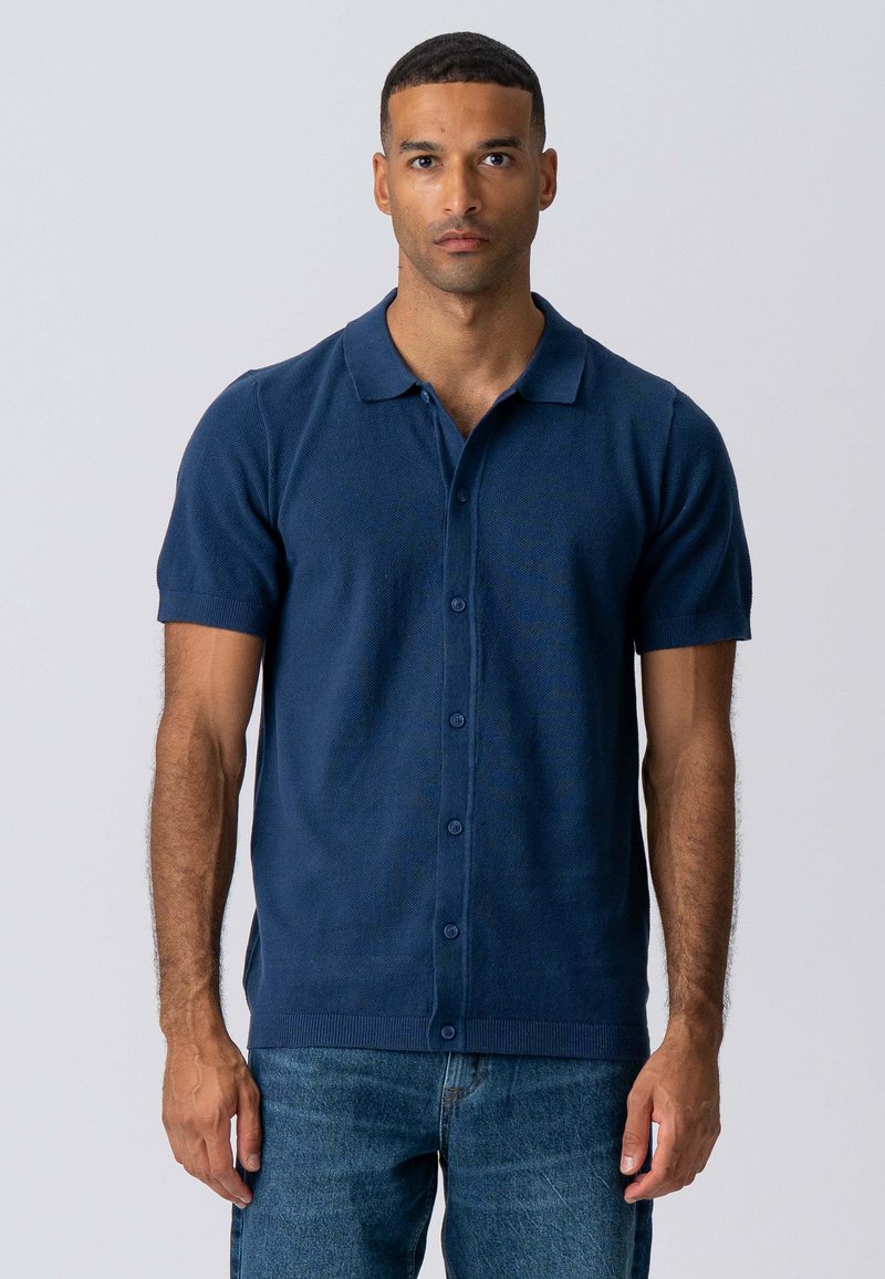 Marineblå kortærmet polo shirt lavet af strukturstof, med knaplukning foran, blød krave og ribbede manchetter, parret med denimjeans.