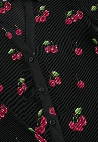 Blouse en tissu noir à côtes avec broderie de cerises roses, mettant en avant des feuilles vertes ; ornée de boutons noirs le long de la patte de boutonnage avant.