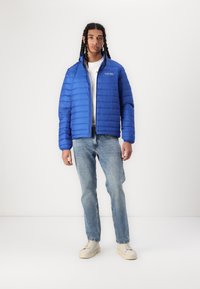 Blaues пухерjacke mit horizontalem gestepptem Design, hohem Kragen und Reißverschluss. Getragen über einem weißen Hemd, kombiniert mit hellblauen Jeans und cremefarbenen Sneakers.