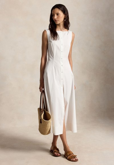 Woman wearing sleeveless white button-front midi dress, holding a woven bag, and wearing brown sandals, standing on a neutral background.