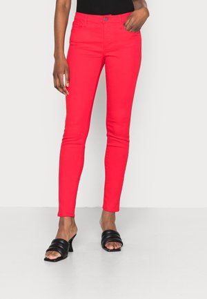 Vrouw draagt felrode slim-fit broek met voorzakken en zwarte open teen sandaal met hak, staand tegen een effen achtergrond.