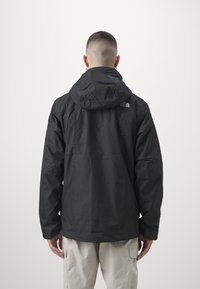 Giacca nera impermeabile con cappuccio, dotata di polsini elasticizzati e un logo sulla schiena. Il modello indossa pantaloni cargo di colore chiaro.