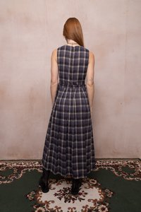 Frau mit langen roten Haaren, die ein ärmelloses, knöchellanges kariertes Kleid trägt, steht auf einem gemusterten Teppich und schaut eine beige Wand an.