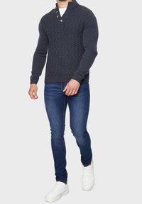 Marineblauwe gebreide trui met een gevarieerd patroon, uitgerust met een sjawl-kraag en drie knopen; gedragen met donkerblauwe jeans en witte sneakers.