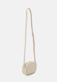 Béžová crossbody kabelka s pevně tvarovaným oválným tvarem, prošívanou texturou a nastavitelným popruhem se zlatým kováním. Horní zapínání na zip.