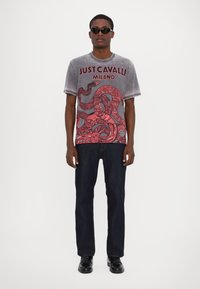 Grijze grafische T-shirt met rood slangdesign, tekst "JUST CAVALLI MILANO". Gecombineerd met donkere spijkerbroek en zwarte schoenen. Zonnebril op.