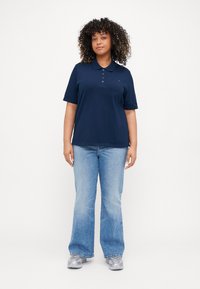 Polo navy con colletto e tre bottoni, abbinato a jeans svasati blu chiaro. Il modello indossa sneakers grigie chiare. Tessuto dal tocco morbido.