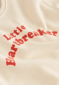 Sahnfarbener Pullover mit der Aufschrift "Little Heartbreaker" in fetten roten Lettern. Weiche Textur mit geripptem Halsausschnitt und lässigem Design.