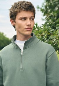 Sudadera de cremallera verde claro hecha de tela suave, con un cuello alto y una cremallera plateada, superpuesta a una camisa blanca.
