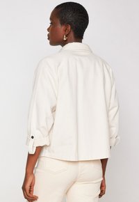 Giacca cropped beige chiaro dalla vestibilità rilassata, maniche arrotolate con dettagli a bottone, e tessuto morbido e testurizzato. Design semplice senza motivi.