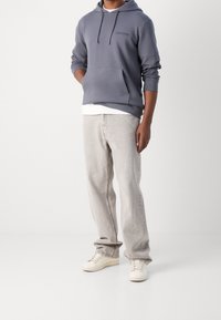 Felpa grigia con tasca a canguro e cordini, indossata sopra una maglietta bianca, abbinata a jeans grigio chiaro e sneaker beige.