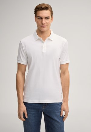 Junger Mann steht frontal und trägt ein schlichtes weißes Polohemd mit kurzen Ärmeln und blaue Jeans vor neutralem Hintergrund.