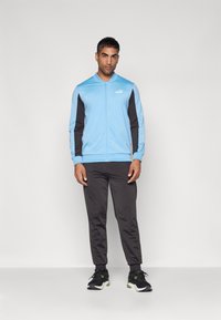 Jachetă cu fermoar de culoare albastru deschis, cu mâneci negre, asortată cu pantaloni negri și adidași negri cu accente verzi. Material moale, sport.