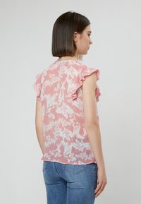 Rosa Bluse mit weißen Blumenmustern, kurzen Rüschenärmeln und einem weichen, leichten Stoff. Kombiniert mit blauen Jeans.