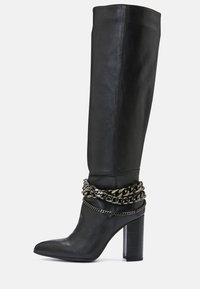 Musta nahkainen polven korkeudelle ulottuva saapas, jossa on teräväkärkinen muotoilu, paksu lohkoheel ja hopeiset ketjuaksentit nilkan ympärillä.