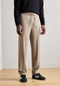 Pantalons beiges ajustés avec une taille élastique et un cordon de serrage. Matériau lisse, coupe ample et jambes droites. Portés avec des baskets noires.